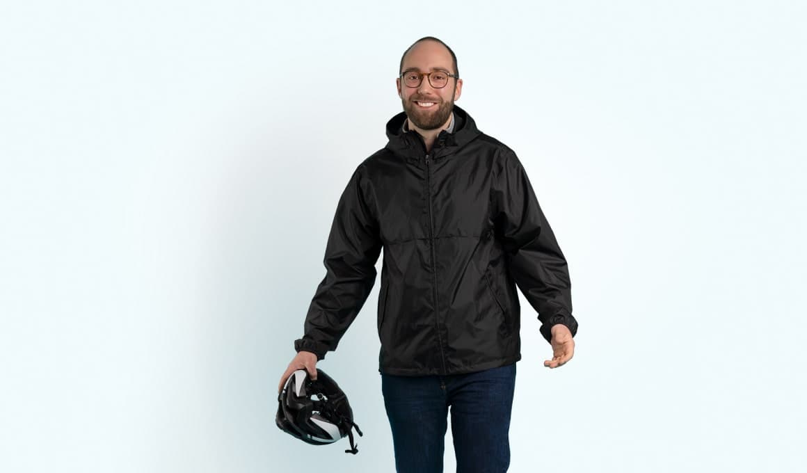 Ein Mann steht in schwarzer Regenjacke gekleidet und hält einen Fahrradhelm in seiner rechten Hand.
