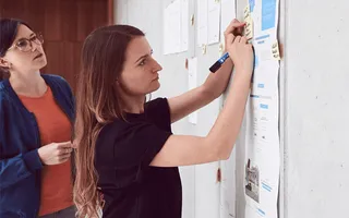 Janette steht am Whiteboard und skizziert, hinter ihr steht ihre Kollegin Maren.