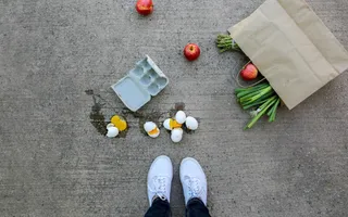 Foto einer heruntergefallenen Papiertüte, aus der Lebensmittel wie Äpfel kullern, daneben eine Packung zerbrochene Hühnereier und die Füße der Person, die das Foto schießt.