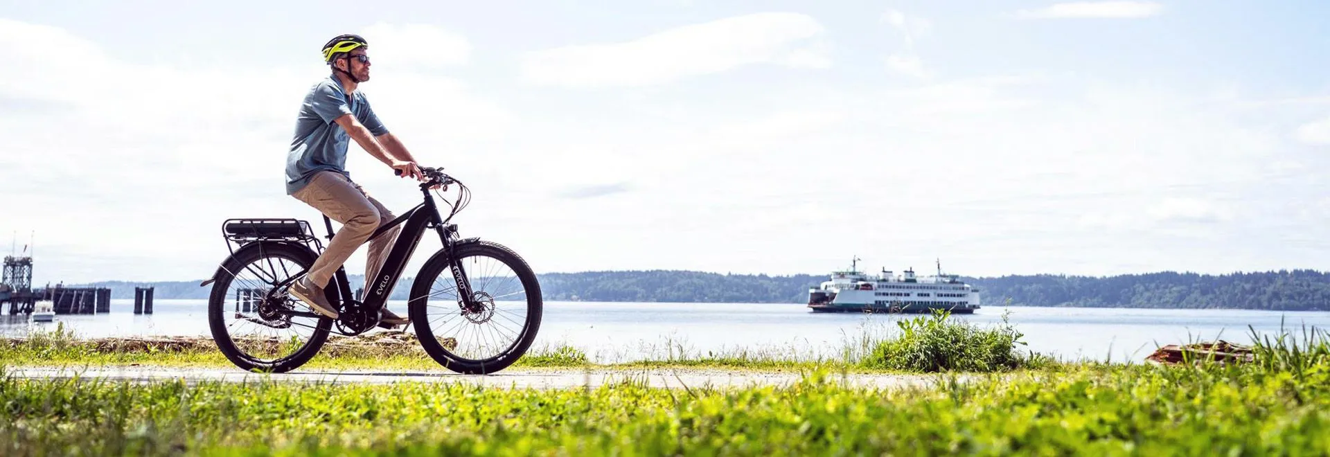 Ein Mann fährt E-Bike auf einer frühlingshaften Wiese entlang eines Flusses. Die Sonne scheint und im Himmel hängen ein paar Schönwetter­wolken. Im Hintergrund sieht man ein Schiff den Fluss hinabfahren.