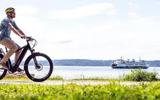 Ein Mann fährt E-Bike auf einer frühlingshaften Wiese entlang eines Flusses. Die Sonne scheint und im Himmel hängen ein paar Schönwetter­wolken. Im Hintergrund sieht man ein Schiff den Fluss hinabfahren.