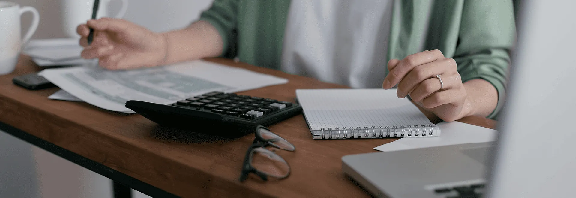 Schufa-Score verbessern geht auch ohne Stift und Papier: Eine Person sitzt vor ihrem Laptop am Schreibtisch und hat Unterlagen, einen Taschenrechner und eine Brille neben sich.