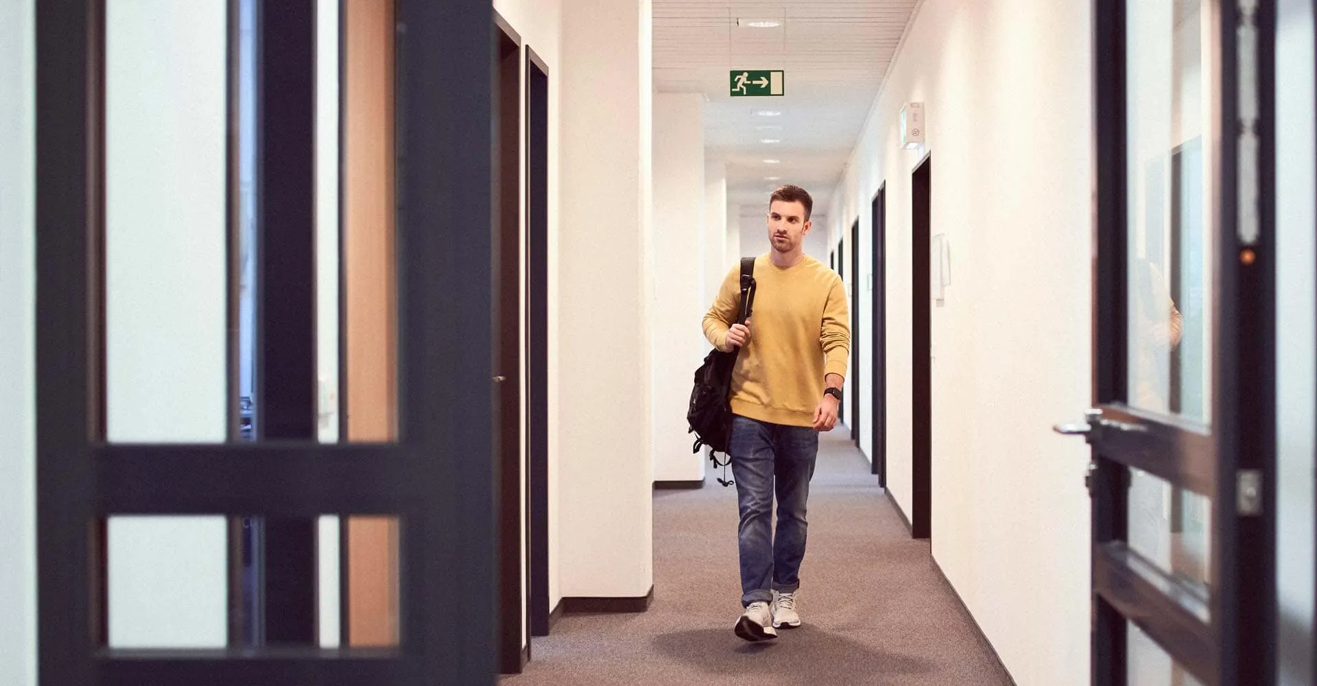 Tobi, wie er im Office ankommt und im gelben Pulli und mit Rucksack den Flur entlang läuft.