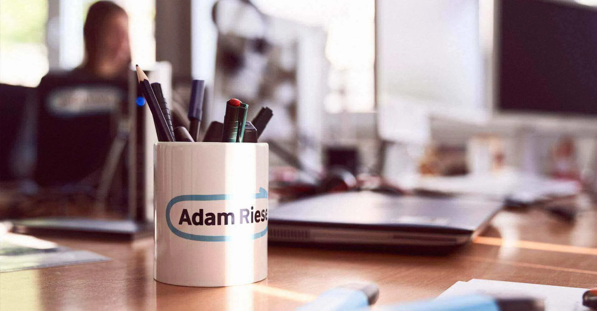 Ein Arbeitsplatz im Office mit Laptops und einer Adam-Riese-Tasse mit bunten Stiften drin